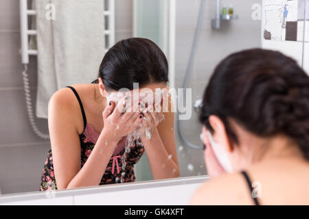 Jeune femme avec un masque cosmétique pulvériser de l'eau sur son visage en face du miroir dans la salle de bains moderne carrelée à la maison Banque D'Images