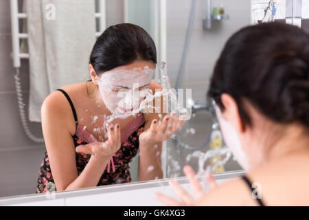 Femme avec masque cosmétique pulvériser de l'eau sur son visage en face du miroir dans la salle de bains moderne carrelée à la maison Banque D'Images