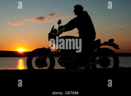 Motocycliste motorbiker en silhouette au coucher du soleil par un lac homme assis sur moto les deux mains au guidon Banque D'Images