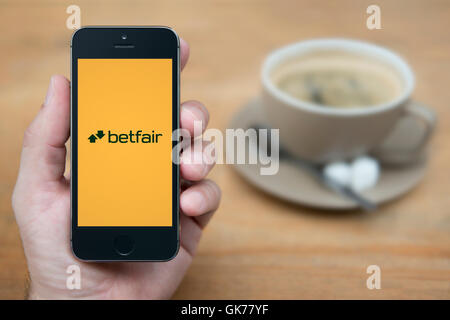 Un homme se penche sur son iPhone qui affiche le logo de Betfair, tandis qu'assis avec une tasse de café (usage éditorial uniquement). Banque D'Images