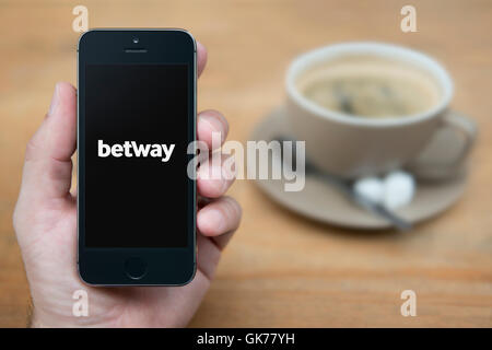 Un homme se penche sur son iPhone qui affiche le logo Betway, tandis qu'assis avec une tasse de café (usage éditorial uniquement). Banque D'Images