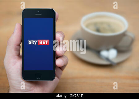 Un homme se penche sur son iPhone qui affiche le logo Sky Bet, alors qu'assis avec une tasse de café (usage éditorial uniquement). Banque D'Images