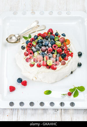 Des Pavlova gâteau avec frais jardin de petits fruits et de cuillères en argent sur blanc sur plaque de fond en bois Banque D'Images