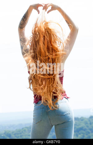 Vue arrière de belle tête rouge en skinny jeans et haut rouge, en montagne au soleil d'or, le vent dans les cheveux et les bras levés. Banque D'Images