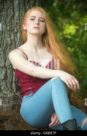 Belle tête rouge femme en skinny jeans et haut rouge, assis sur des aiguilles de pin avec en arrière contre un arbre.