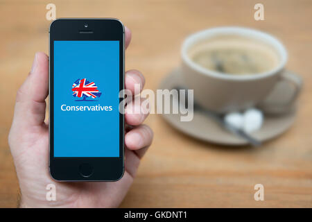 Un homme se penche sur son iPhone qui affiche le logo des conservateurs, alors qu'assis avec une tasse de café (usage éditorial uniquement). Banque D'Images