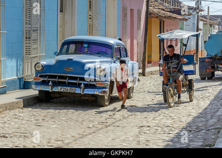 Scène de rue avec une voiture américaine classique, un vélo-taxi et un jeune garçon courir à Trinité, la province de Sancti Spiritus, Cuba Banque D'Images