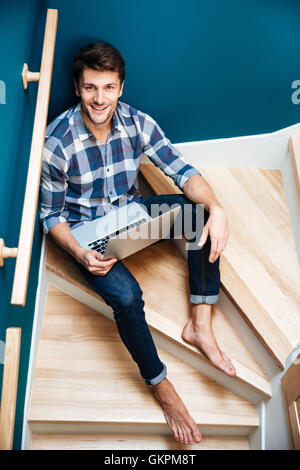 Vue de dessus de joyeux jeune homme en chemise à carreaux assis sur des escaliers à la maison et à l'aide d'ordinateur portable Banque D'Images