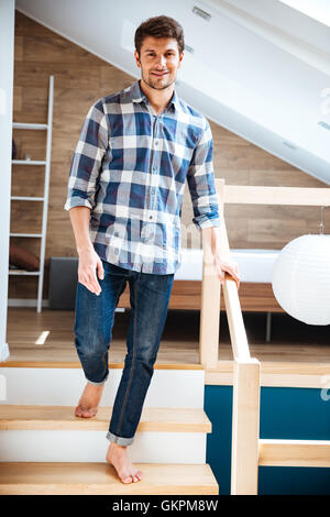 Beau jeune homme en jeans et chemise à carreaux marche pieds nus sur des escaliers à la maison Banque D'Images