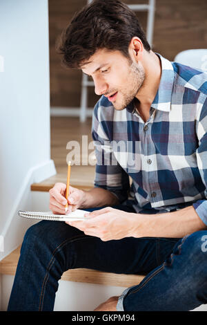 Jeune homme concentré en chemise à carreaux assis et l'écriture dans le bloc-notes à la maison Banque D'Images