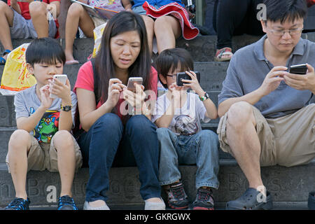 Une famille asiatique assis sur les marches à la bibliothèque publique au centre-ville de Flushing, Queens, NYC, tous à l'aide de leur téléphone cellulaire. Banque D'Images