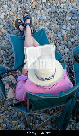 Hauts femme lecture livre assis dans le siège à shingle beach Banque D'Images