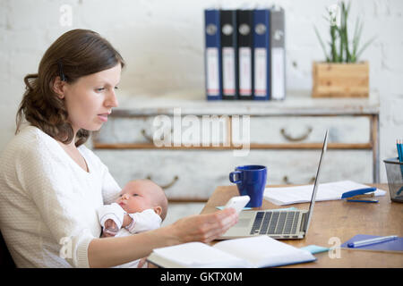 Portrait of young business maman tenant son nouveau-né bébé mignon tout en travaillant à l'intérieur du home office, en regardant l'écran du téléphone cellulaire Banque D'Images