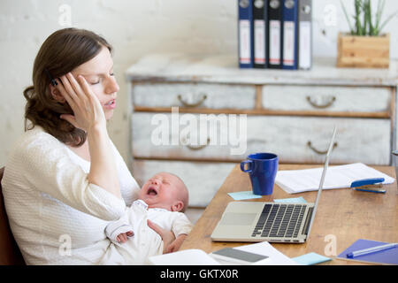 Young businesswoman holding newborn bébé pleurer alors qu'il était assis à l'ordinateur portable et de toucher la tête avec expression douloureuse. Maman de carrière Banque D'Images