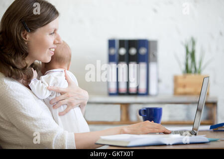 Vue de côté portrait of happy casual young business maman tenant son bébé mignon nouveau-né tandis que working on laptop in home office inter Banque D'Images