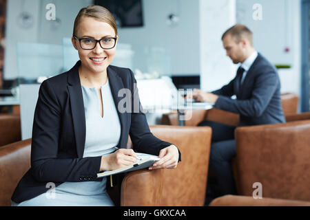 Happy young businesswoman looking at camera while making notes dans l'ordinateur portable Banque D'Images