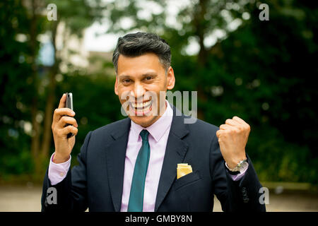 Homme d'affaires joyeuse à peau d'olive avec bras levés et tenant le téléphone mobile dans une main. Banque D'Images