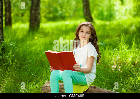 Charmante petite fille en forêt avec livre sur l'arrière-plan de parc d'été Banque D'Images