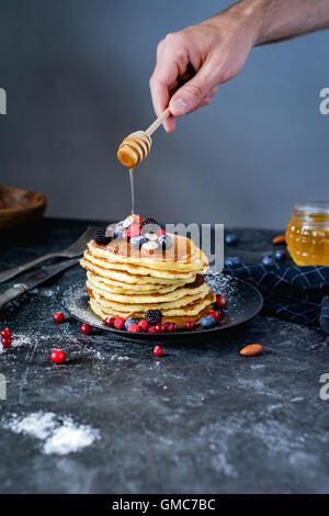 Des crêpes pour le petit-déjeuner. Homme main tenant une cuillère de miel et verser le miel sur pile de crêpes aux fruits d'été et d'écrous. Banque D'Images