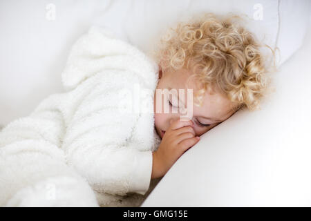 Cute blonde curly petit garçon endormi sur la table blanche. Tout-petits fatigués prendre une sieste portant des pyjamas blanc chaud, suçant son pouce. Banque D'Images