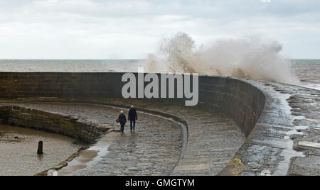 Deux personnes marchant le long de la Cobb à Lyme Regis Dorset sur la côte jurassique, au cours d'une tempête avec des vagues se brisant sur eux. Banque D'Images
