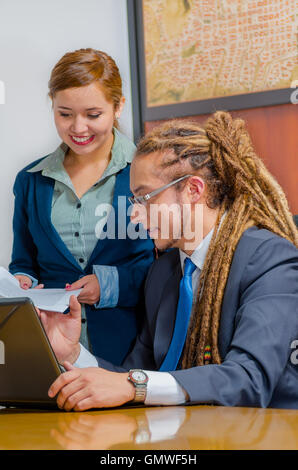 Beau jeune homme portant un costume formel de s'asseoir avec la jolie femme collègue debout à côté de lui à la recherche à l'écran et discuter entre eux, gestionnaire de bureau concept Banque D'Images