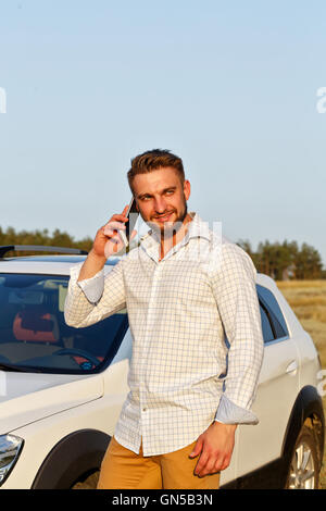 Beau jeune homme debout en face de la voiture. Un homme parle sur un smartphone et sourit. Toujours connecté. Banque D'Images