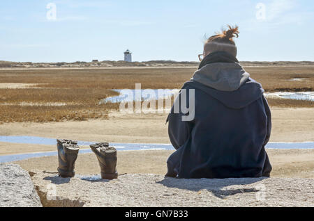 Une jeune femme est assise sur un rocher jetée avec ses bottes à côté d'elle et d'un phare au loin. Banque D'Images