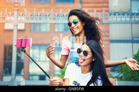 Paire de happy friends making faces tout en prenant un autoportrait à l'extérieur avec un téléphone cellulaire rose attaché à pôle stick selfies Banque D'Images