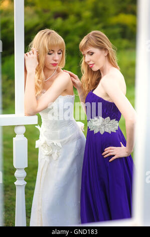 Mariée de demoiselle d'honneur et de l'extérieur. Deux jeunes filles posant sur le pré vert. Mariée en robe de mariage. Dans demoiselle d'une robe de soirée pourpre. Banque D'Images