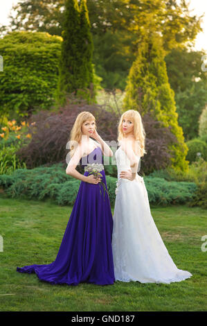 Mariée de demoiselle d'honneur et de l'extérieur. Deux jeunes filles posant sur le pré vert. Mariée en robe de mariage. Dans demoiselle d'une robe de soirée pourpre. Banque D'Images