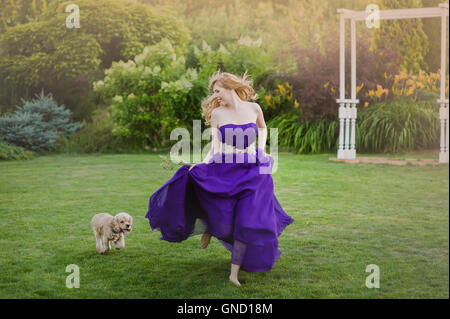 Belle fille courir sur la pelouse avec chien. Dans demoiselle d'une robe de soirée pourpre. Banque D'Images