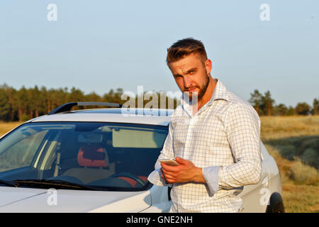 Un beau jeune homme qui se tient devant la voiture. Un homme tenant un smartphone. Toujours connecté. Banque D'Images