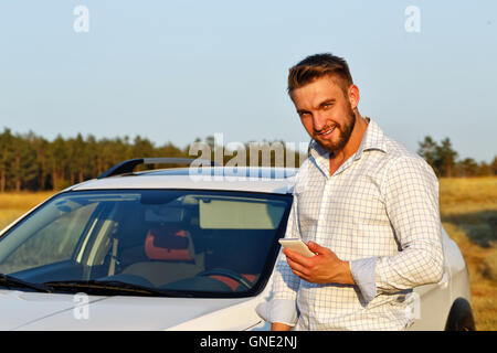 Un beau jeune homme qui se tient devant la voiture. Un homme tenant un smartphone et sourit. Toujours connecté. Banque D'Images