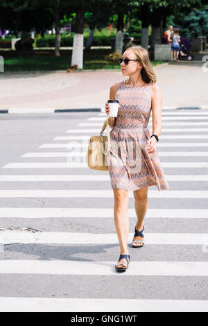 Jeune brunette woman in dress traversant la rue avec Coffee cup Banque D'Images