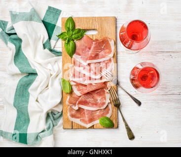 Tranches de jambon prosciutto di Parma et feuilles de basilic frais sur planche de bois et verres à vin rose sur fond en bois laqué blanc Banque D'Images
