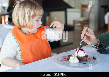 Deux ans enfant de manger avec orange cuillère en plastique un morceau de gâteau au chocolat avec crème glacée à la vanille partager avec femme à restau Banque D'Images