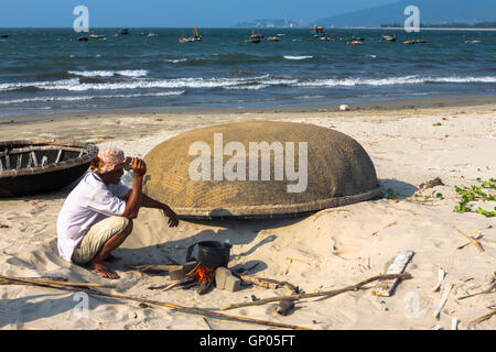 Da Nang Bay : pêcheur tend son feu sur la plage, avec les coracles (-chai thung) au-delà, Da Nang, Viet Nam Banque D'Images