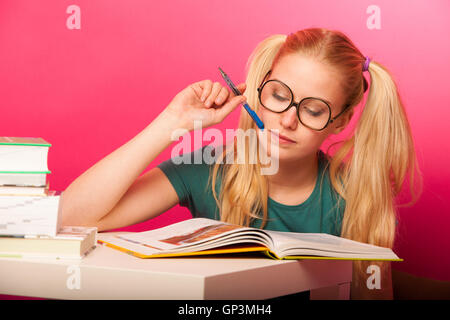 Écolière espiègle avec de grandes lunettes concentré dans la lecture de livre assis sur le plancher derrière la petite table. Banque D'Images