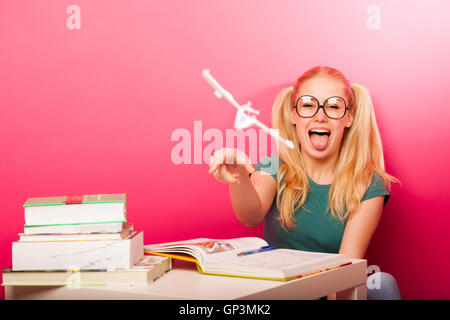 Espiègle, naughty schoolgirl avec de grandes lunettes jouant avec avion en papier alors que devrait étudier. Assis derrière tabe plein de boo Banque D'Images