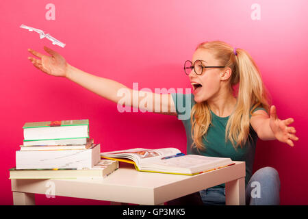 Espiègle, naughty schoolgirl avec de grandes lunettes jouant avec avion en papier alors que devrait étudier. Assis derrière tabe plein de boo Banque D'Images