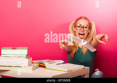 Espiègle, naughty schoolgirl avec de grandes lunettes jouant avec avion en papier alors que devrait étudier. Assis derrière tabe plein de boo Banque D'Images