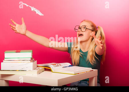 Espiègle, naughty schoolgirl avec de grandes lunettes jouant avec avion en papier alors que devrait étudier. Assis derrière tabe plein de boo Banque D'Images