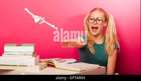 Espiègle, naughty schoolgirl avec de grandes lunettes jouant avec avion en papier alors que devrait étudier. Assis derrière tabe plein de boo Banque D'Images