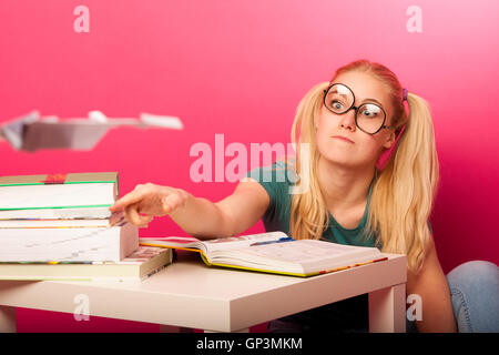 Espiègle, naughty schoolgirl avec de grandes lunettes jouant avec avion en papier alors que devrait étudier. Assis derrière tabe plein de boo Banque D'Images