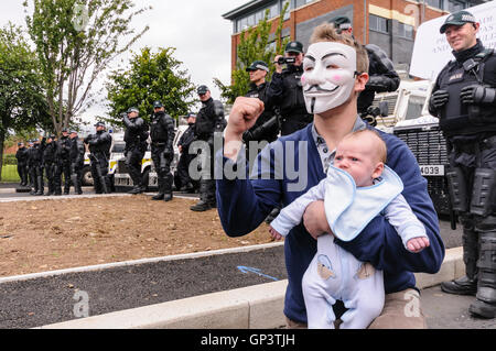 Un homme portant un Guy Fawkes 'Anonyme' masque, et tenant un bébé, donne un air de défi salue en face d'une rangée d'agents de police PSNI Banque D'Images