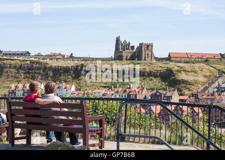 Un homme et une femme assis sur un banc en admirant le port et à l'Est des falaises de Whitby, North Yorkshire y compris l'abbaye Banque D'Images