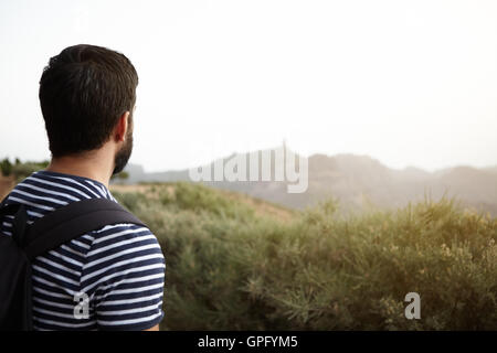 Jeune homme à la recherche sur la montagne en plein soleil portant un t-shirt à rayures avec un sac à dos et une barbe Banque D'Images