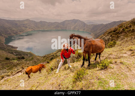 Femme blonde avec deux chevaux près du bord de la montagne, lac de Quilotoa en arrière-plan, l'Équateur, en Amérique du Sud Banque D'Images