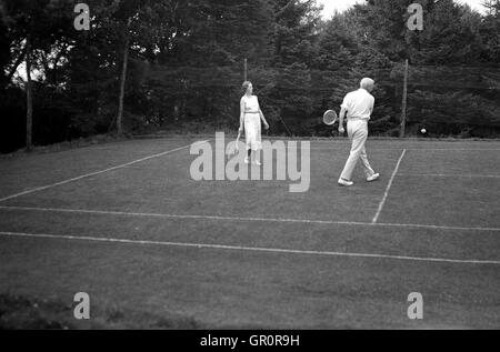 Années 1930, historiques, en lecture sur un couple à l'extérieur de tennis sur gazon, Spreyton village, Devon, Angleterre. Banque D'Images